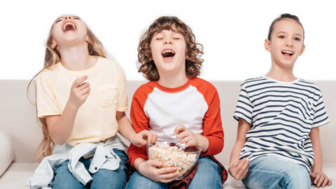 3 children laughing on a couch, with bowl of popcorn