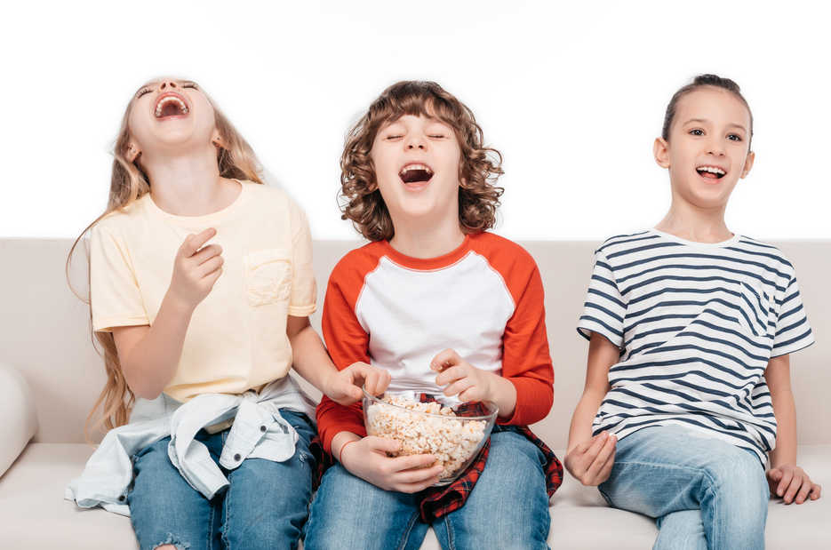 3 children laughing on a couch, with bowl of popcorn